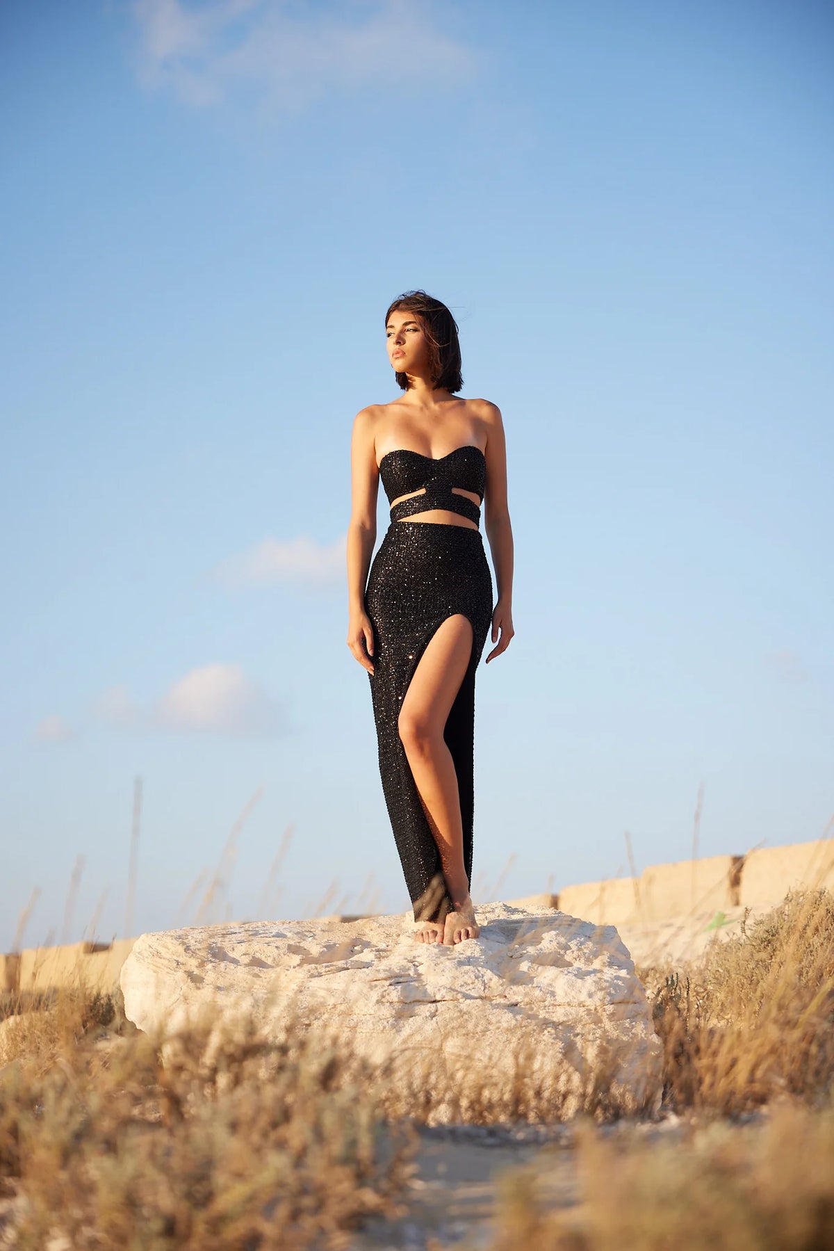 A woman standing on a rock wearing a black sequin embellished top and maxi skirt with a high slit, posing against a backdrop of dry grass and sky.