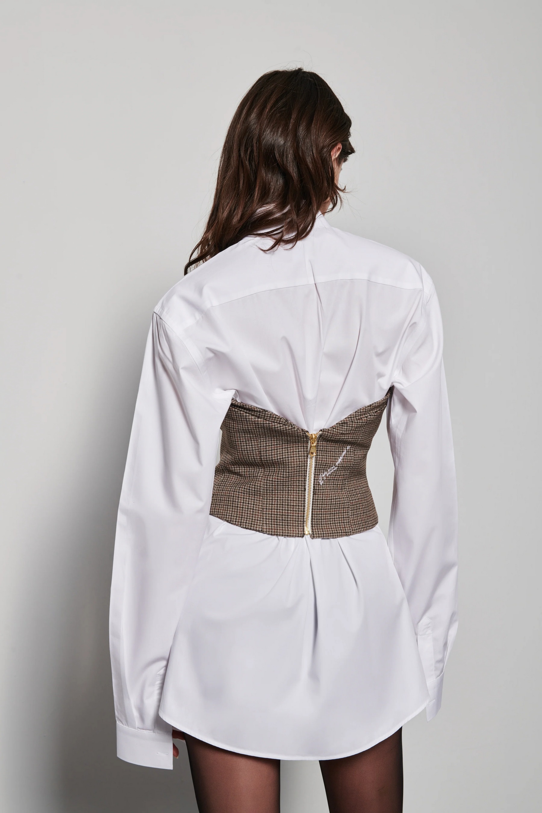 A model wearing a brown checkered bustier with collar details and golden buttons, on top of a white chemise dress