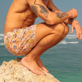 Man with tattoos wearing colorful swim trunks sitting on a rock by the ocean.