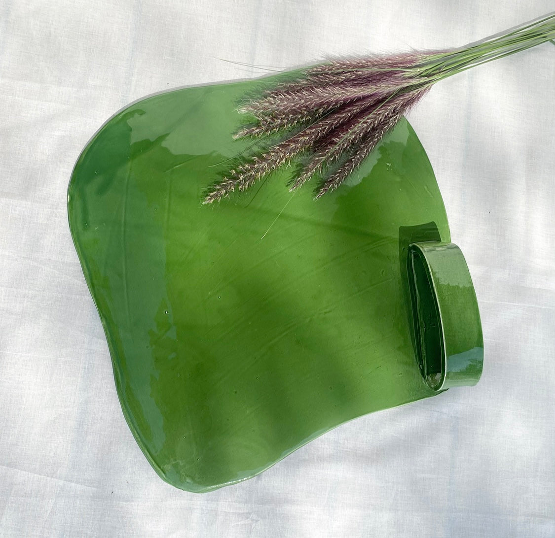 A green ceramic cheese platter with a handle, displayed on a white surface with a sprig of dried flowers placed on top for decoration.