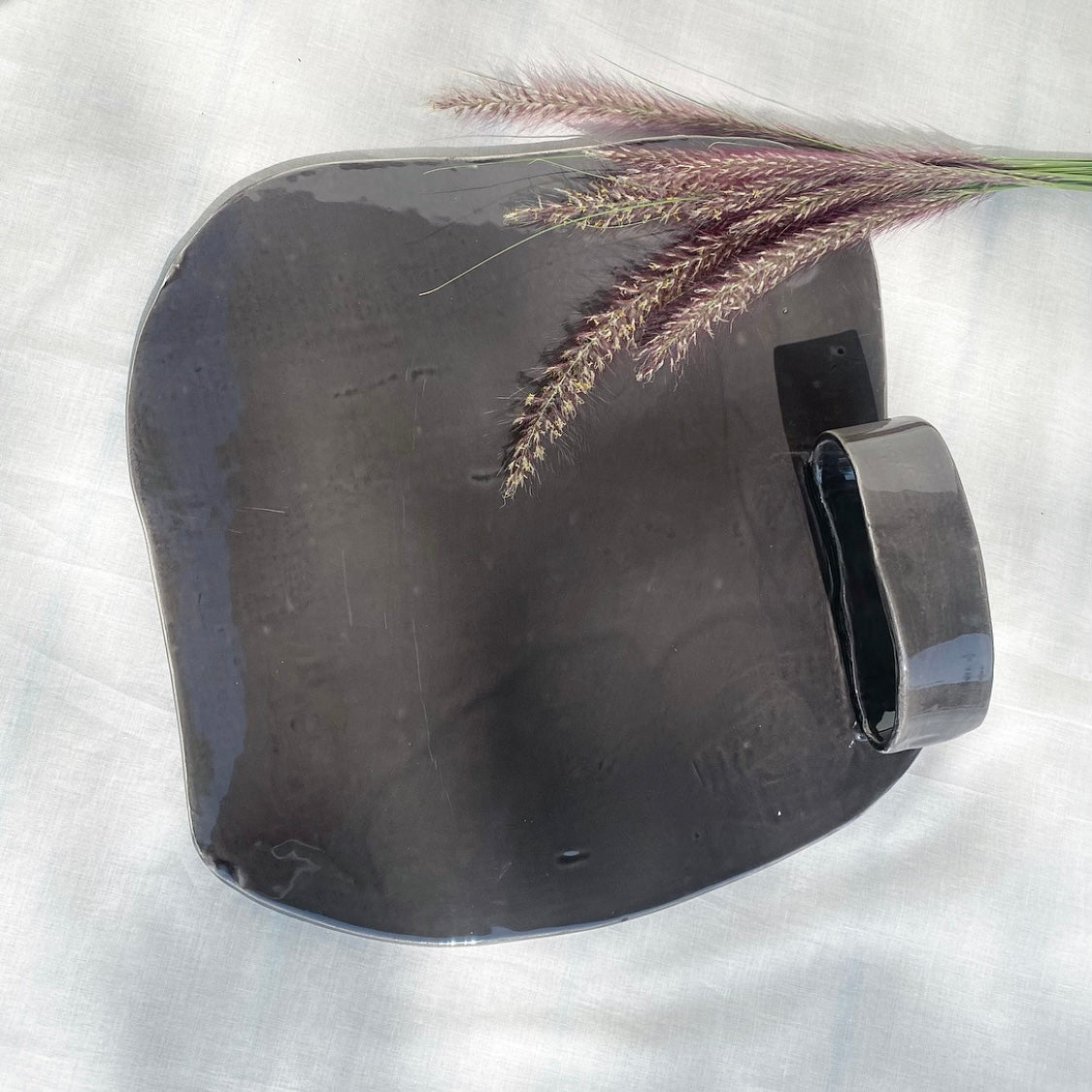 A gray ceramic cheese platter with a handle, displayed on a white surface with a sprig of dried flowers placed on top for decoration.