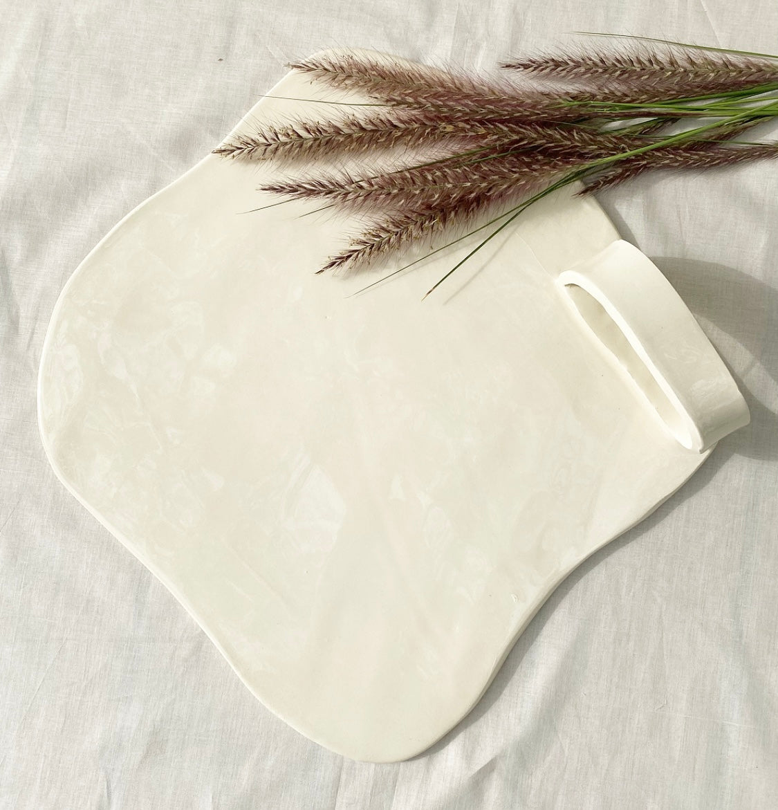 A white ceramic cheese platter with a handle, displayed on a white surface with a sprig of dried flowers placed on top for decoration.