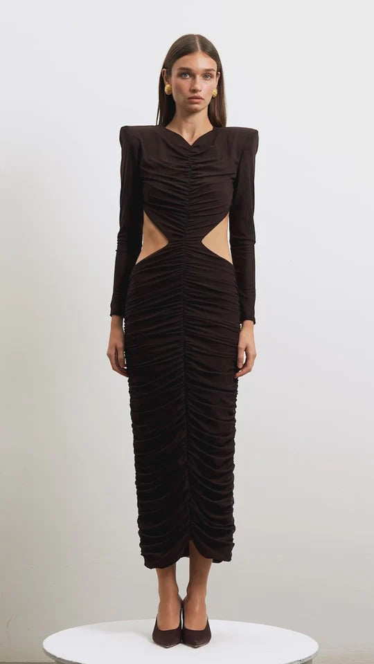 A woman standing and facing to the side wearing a brown maxi dress with a cut-out back and a decorative belt.