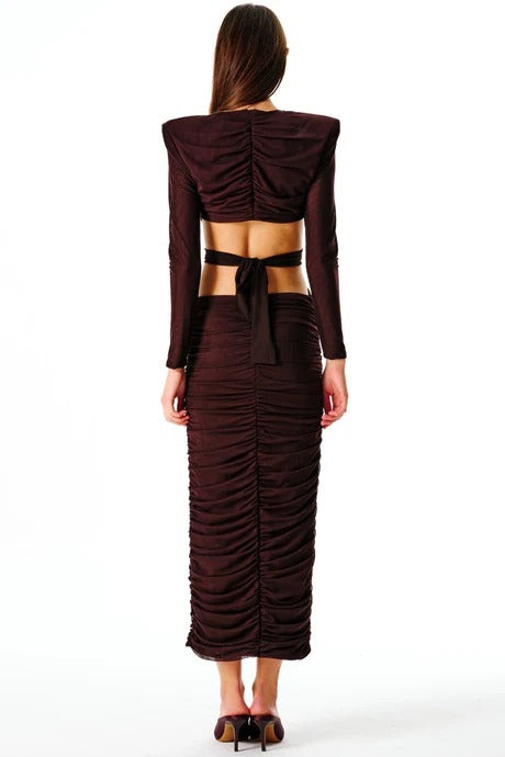 A woman standing and facing to the side wearing a brown maxi dress with a cut-out back and a decorative belt.