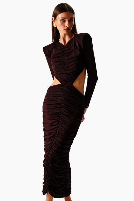 A woman standing and facing to the side wearing a brown maxi dress with a cut-out back and a decorative belt.
