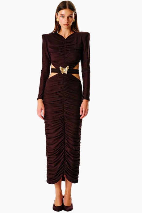 A woman standing and facing to the side wearing a brown maxi dress with a cut-out back and a decorative belt.