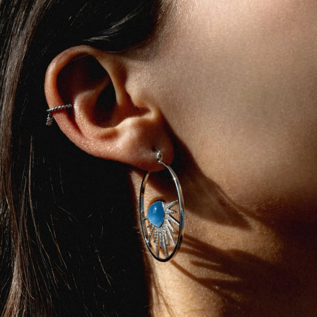 Close-up of an ear wearing a silver hoop earring with a blue gemstone.