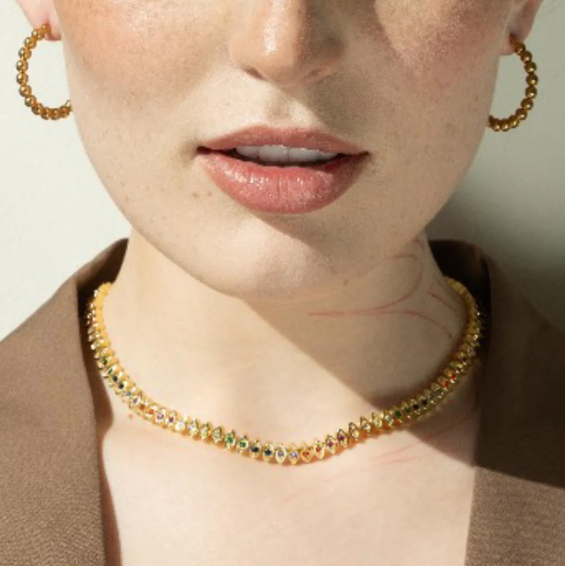 Close-up of a person wearing a gold necklace and hoop earrings with a neutral background.