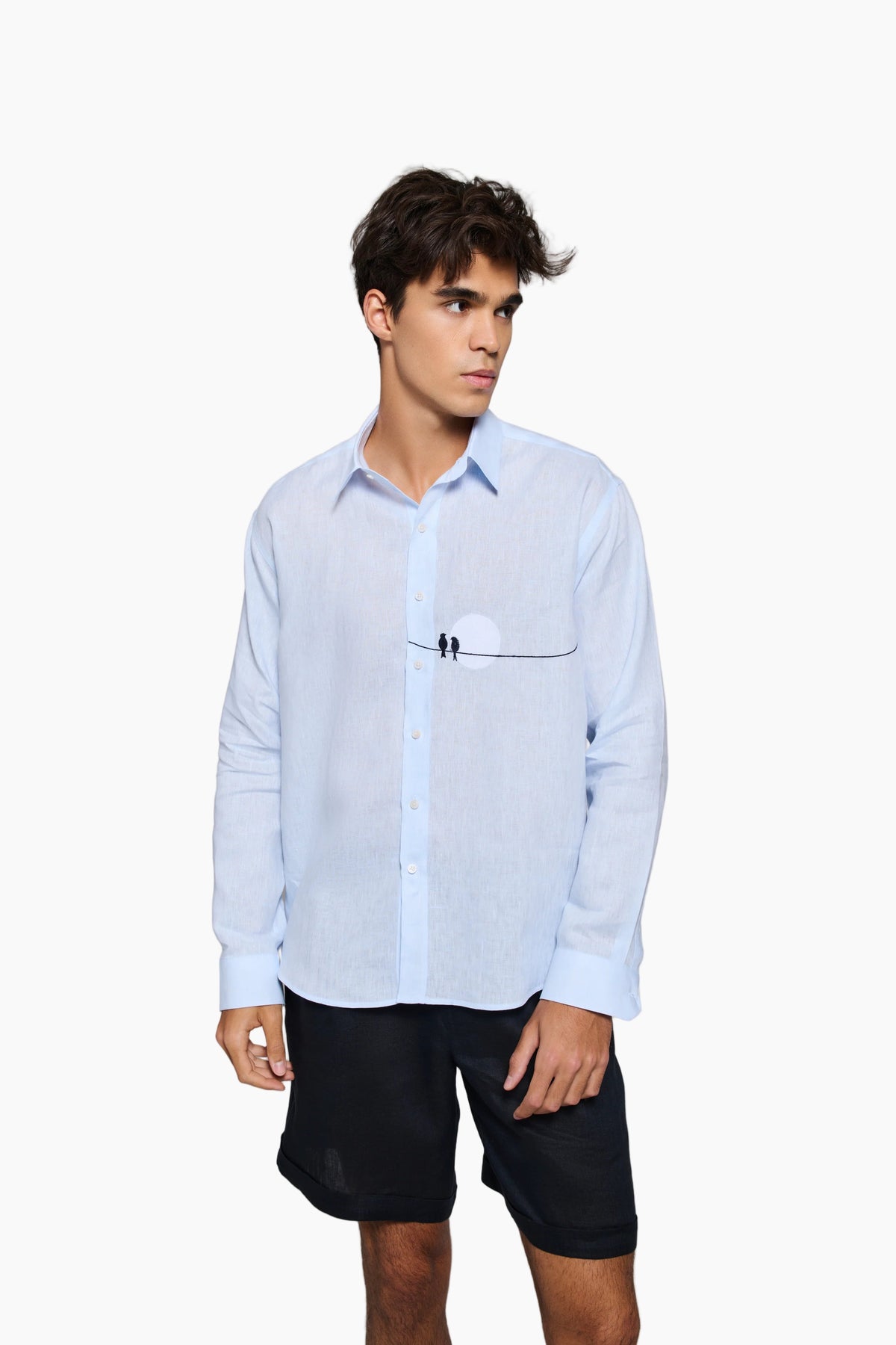 Man wearing a light blue shirt and black shorts : Tailored fit baby blue 100% fine Linen men shirt with a hanging two love birds and a sunset view embroidery on the chest.