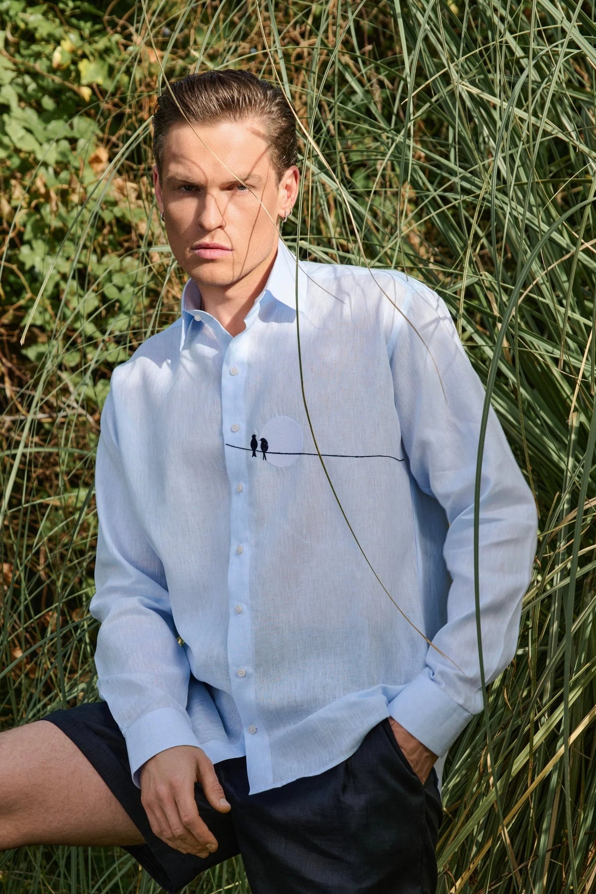 Man wearing a light blue shirt and black shorts : Tailored fit baby blue 100% fine Linen men shirt with a hanging two love birds and a sunset view embroidery on the chest.