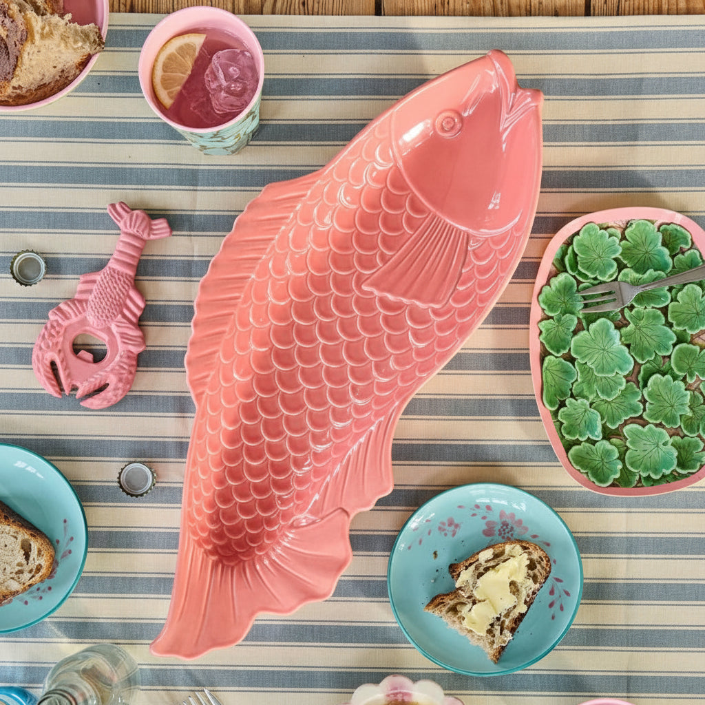 Pink fish-shaped platter on a striped tablecloth with various dishes and utensils.
