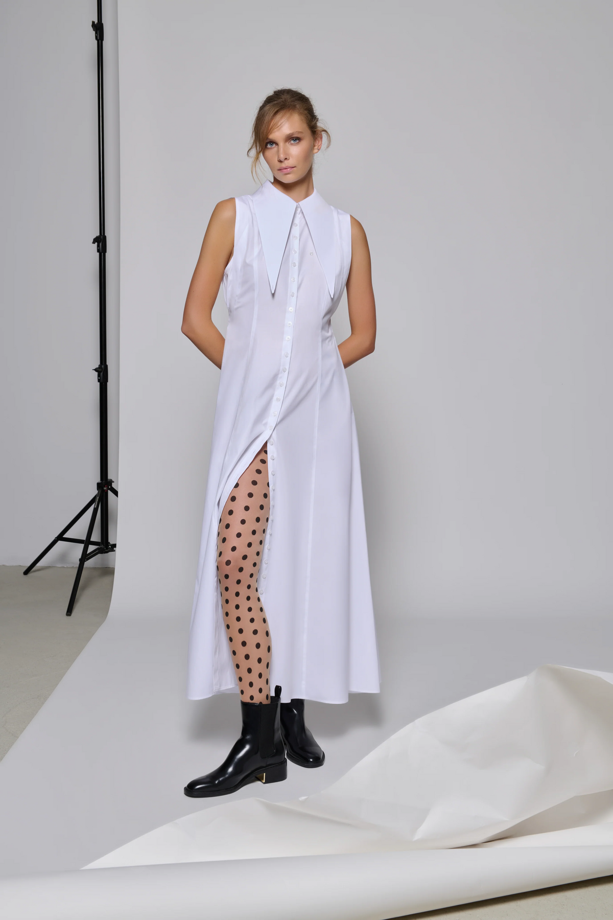 women wearing a long buttoned up White Poplin dress with drapes on the waist and long collars adjustable in wearing methods. The dress has a detachable black tie on the collar and tucked on the back side of the garment.