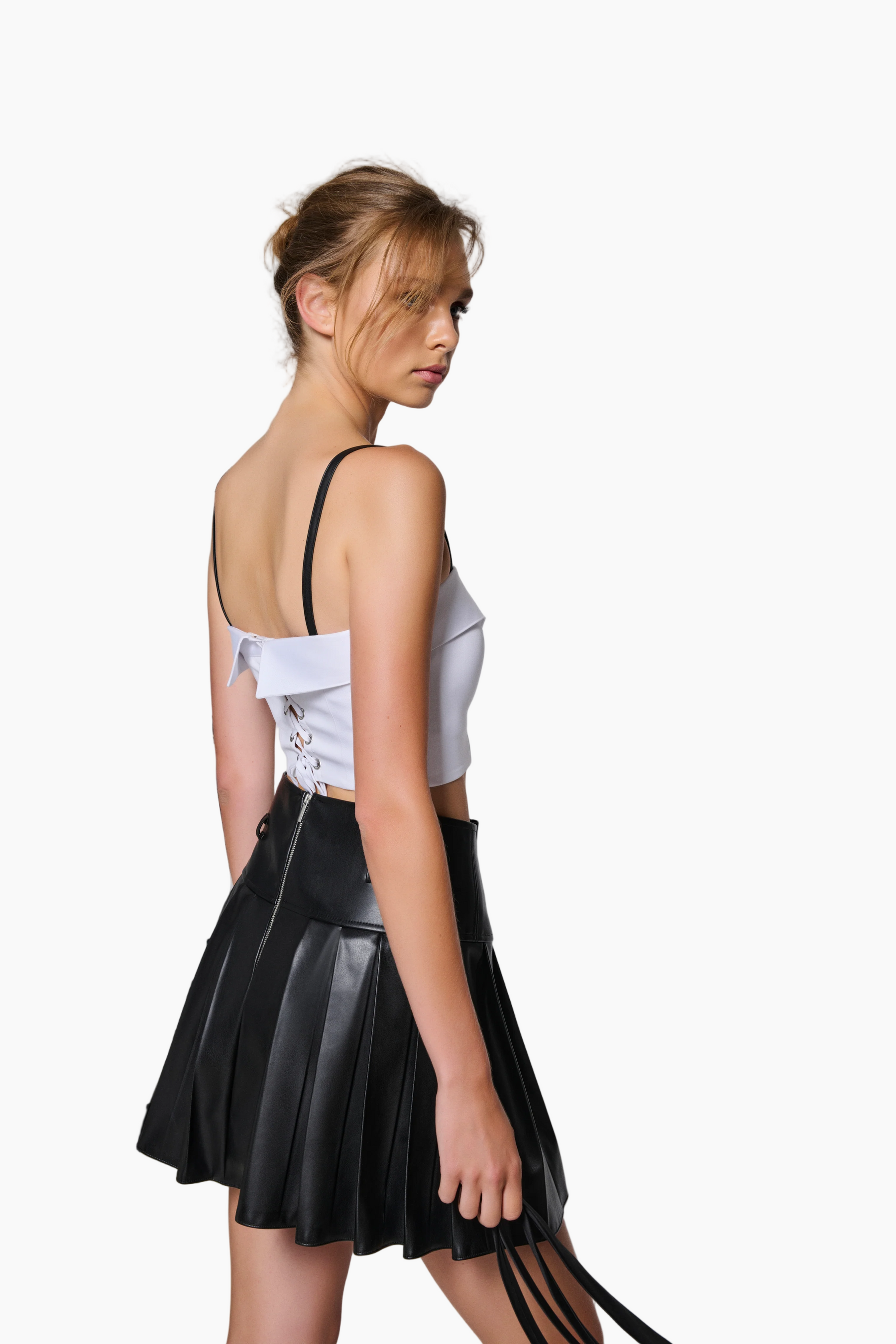 Woman wearing a white top with lace-up back and a black pleated skirt against a plain background