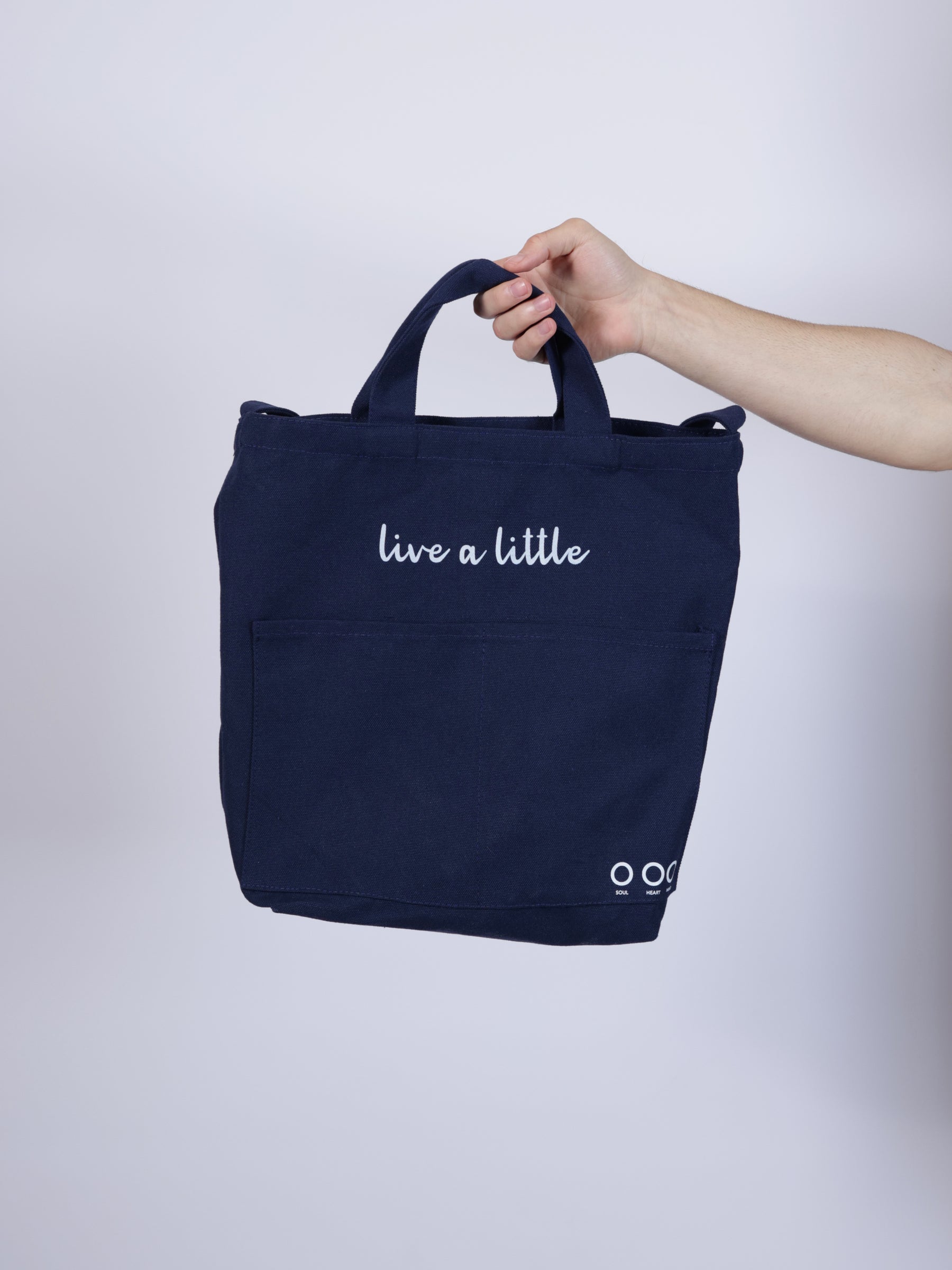 Person holding a navy blue tote bag with 'live a little' text