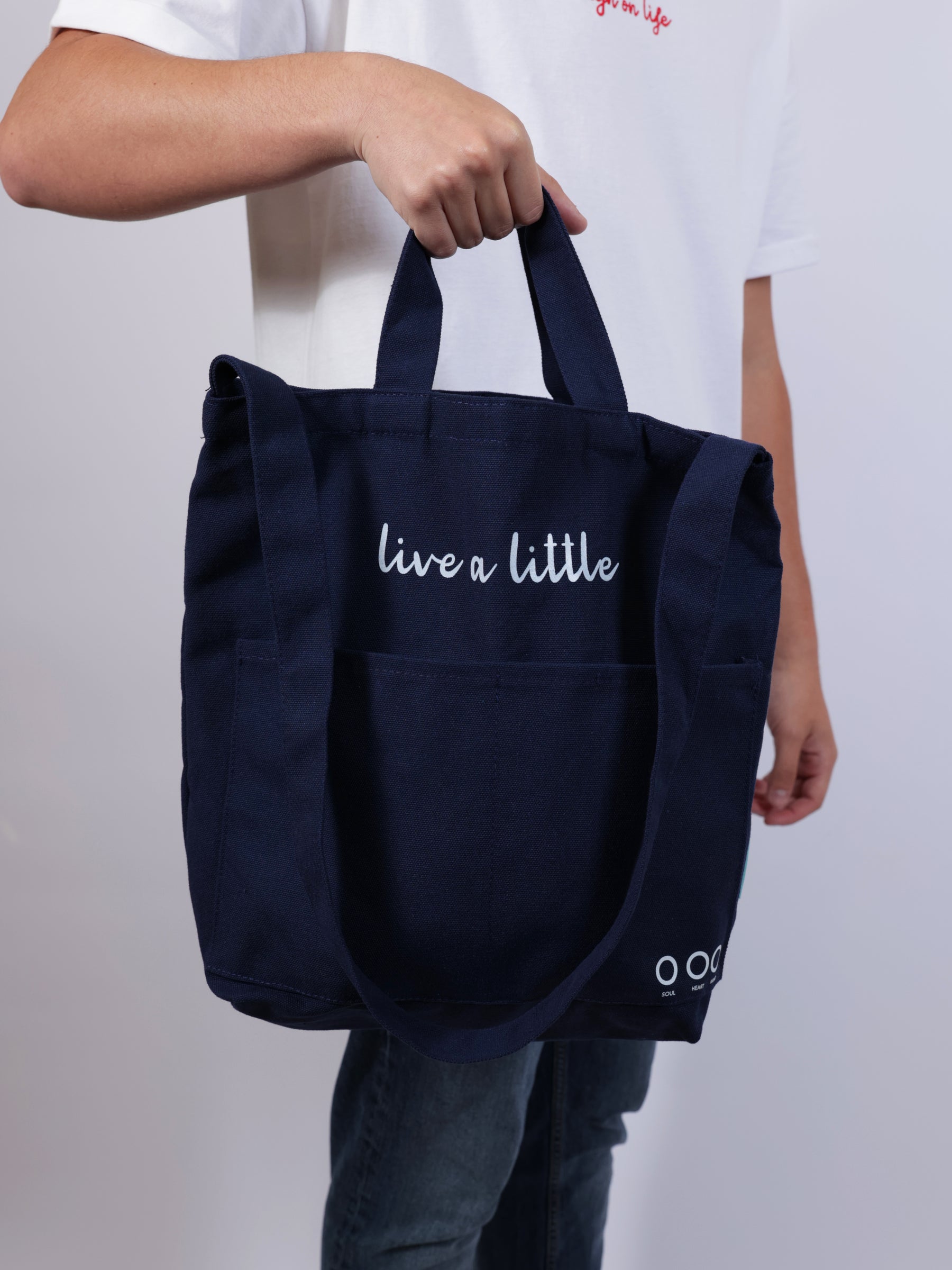 Person holding a navy blue tote bag with 'live a little' text