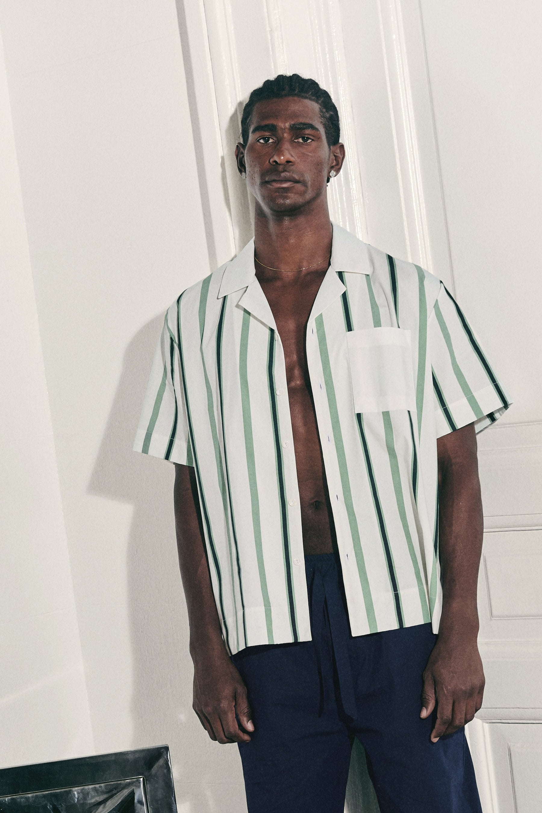 Man wearing a striped shirt and dark pants standing next to a chair against a white paneled wall.