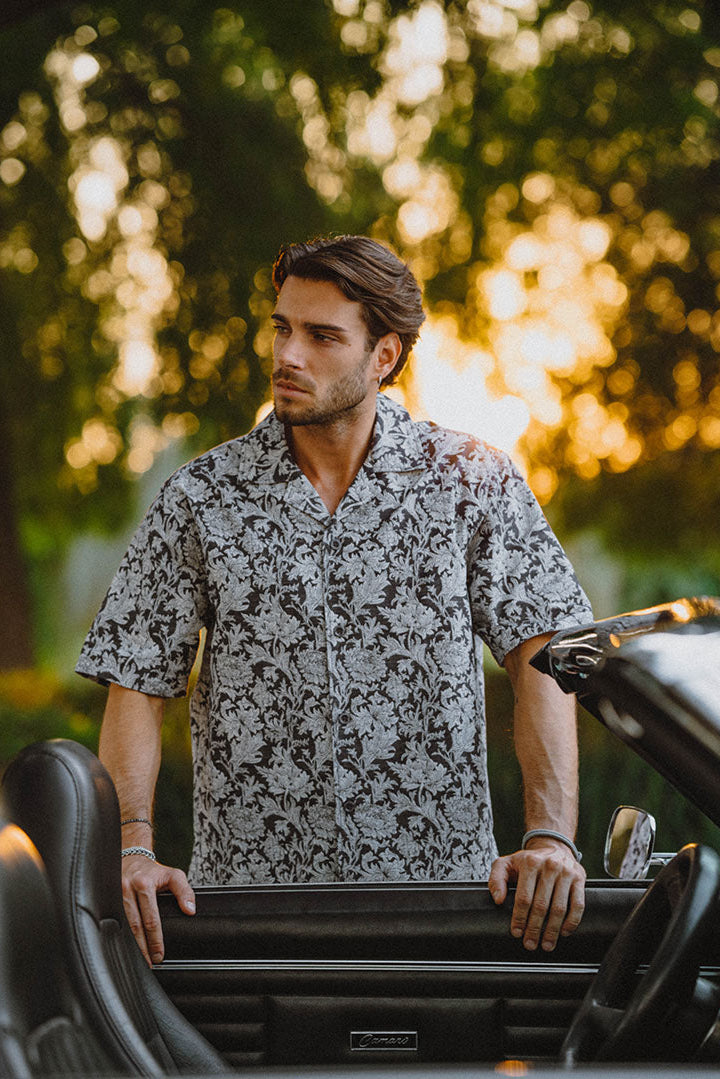 Man standing next to a car with a patterned shirt in light blue and navy.