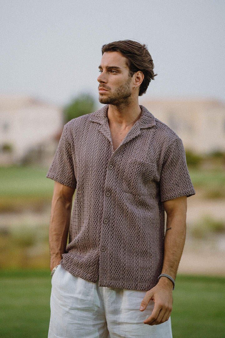 Man wearing a patterned brown shirt and white pants standing outdoors.