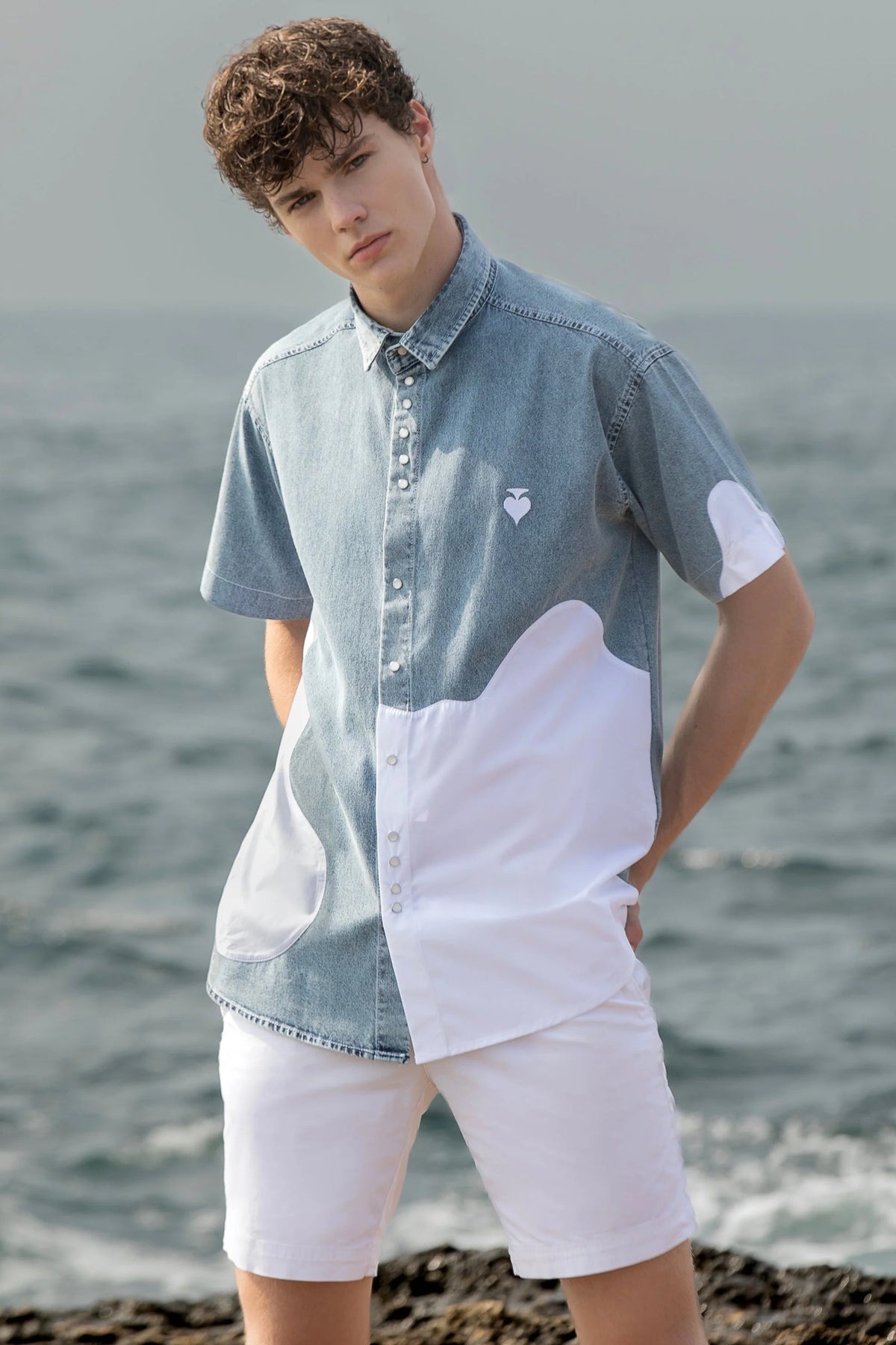 Person wearing a denim and white shirt with a heart design by the ocean. Unisex standard fit half denim - half poplin short sleeve shirt with a handmade spade embroidery on the chest