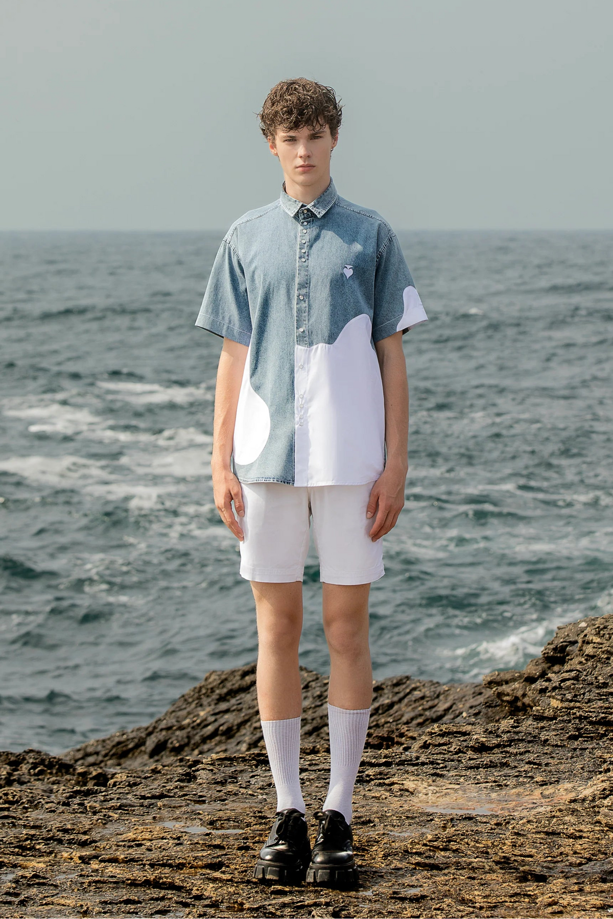 Person standing on a rocky shore wearing Unisex standard fit half denim - half poplin short sleeve shirt with a handmade spade embroidery on the chest