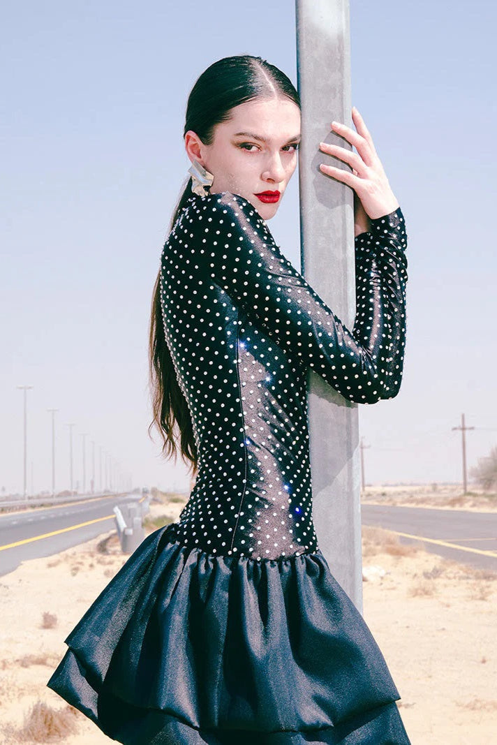 Woman in a black polka dot dress standing by a road with a clear sky.