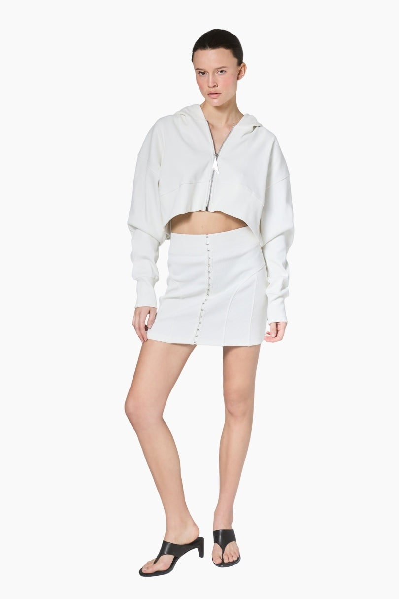 a white cropped hoodie featuring a structured silhouette with dropped shoulders, ribbed texture, and a statement front zipper pull paired with a short white skirt.