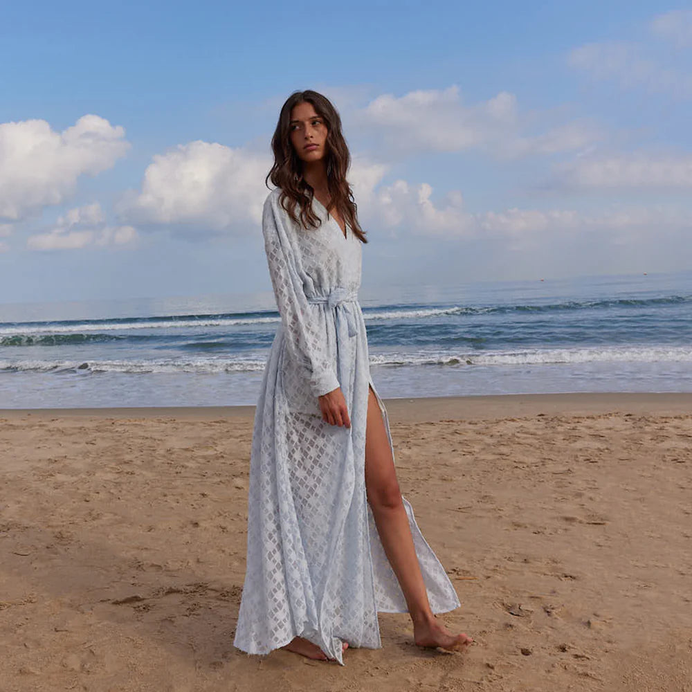 Woman wearing a light blue patterned dress with a slit. Flared floor-length dress with a V-neckline, balloon sleeves, two side slits, and a waist belt.
