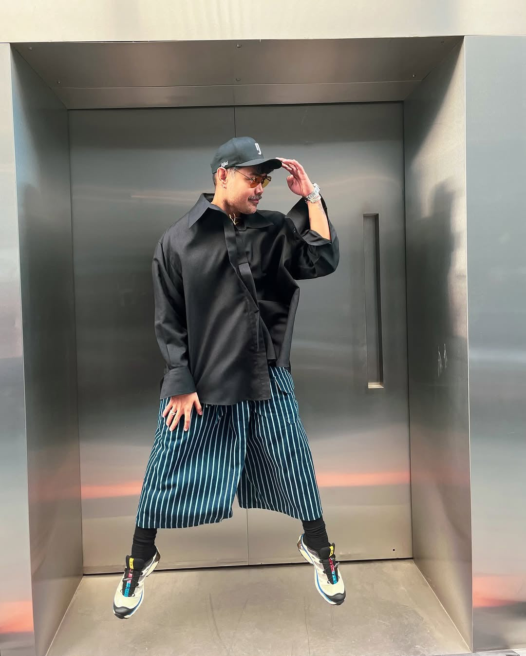 A person standing in front of an elevator wearing fisherman pants with a striped pattern and a black shirt.