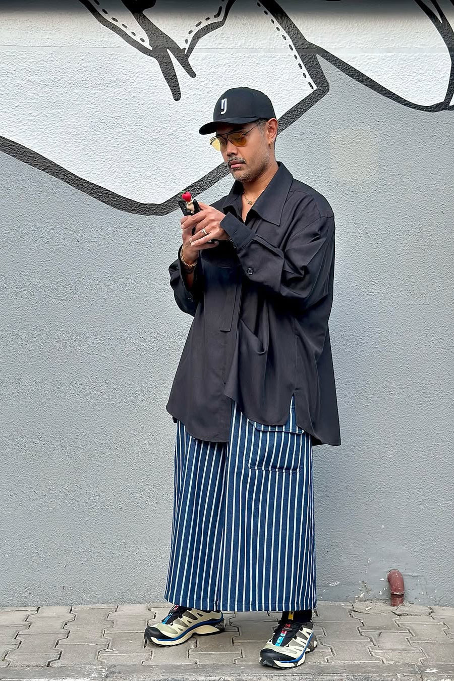 Person wearing a black jacket and striped pants standing against a graffiti-covered wall.