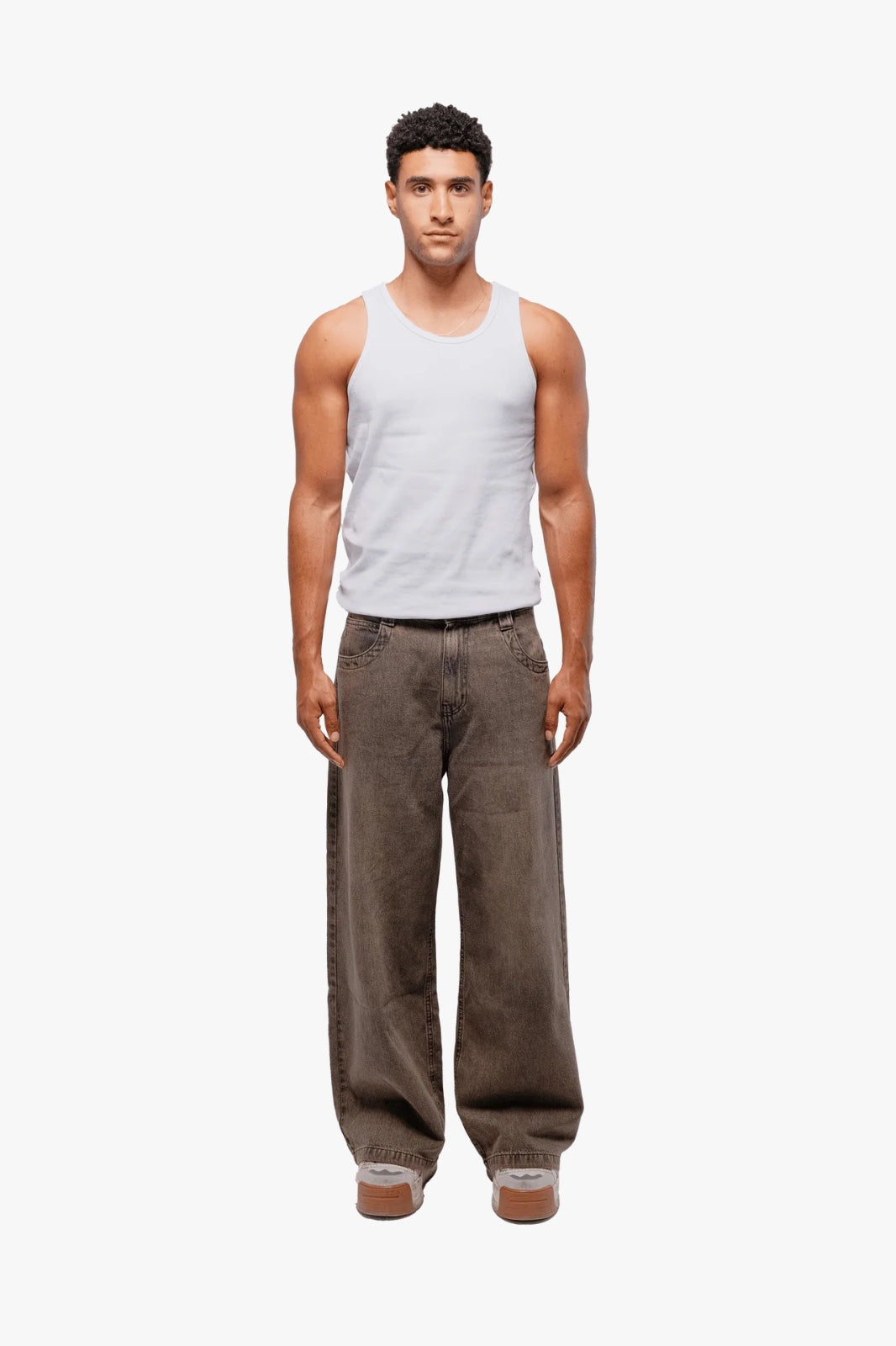 A person standing against a white background, wearing a white tank top and brown baggy pants with multiple pockets.