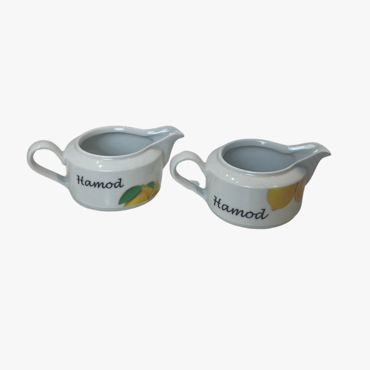 Two white porcelain creamers with the text 'Hamod' written on them, one featuring a lemon illustration and the other an orange illustration, both with a blue background.