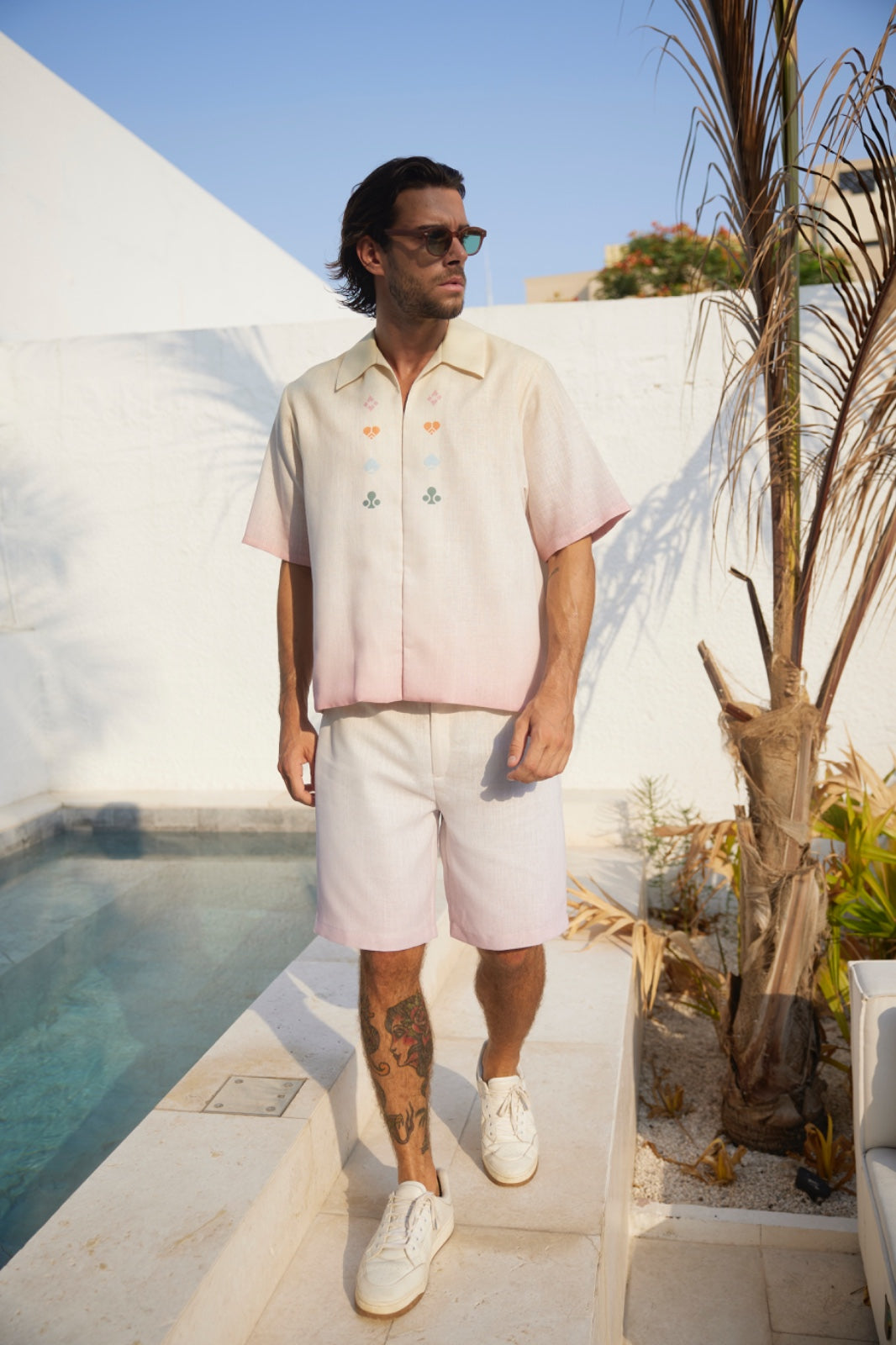 A man standing poolside wearing a white shirt with a key design, white shorts, and white sneakers.