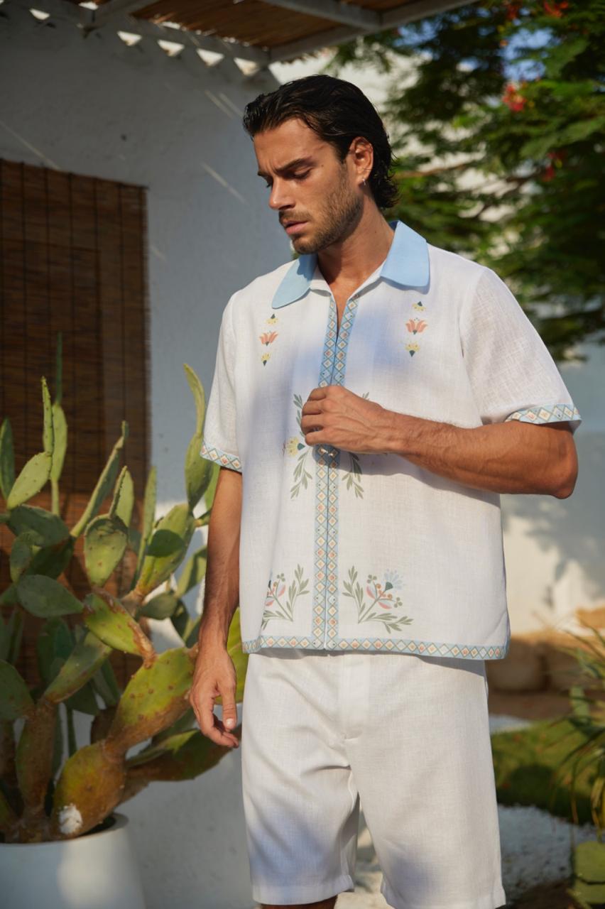 A man wearing a white shirt with blue and multicolored floral embroidery and a collar, paired with white shorts.