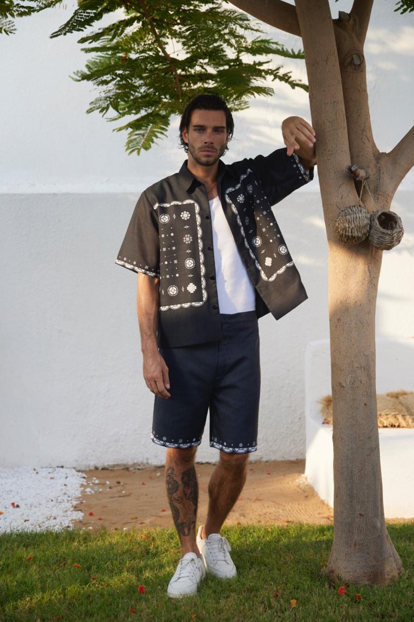 A man standing outdoors wearing a black shirt with a round neckline and short sleeves, paired with dark blue denim shorts and white sneakers.
