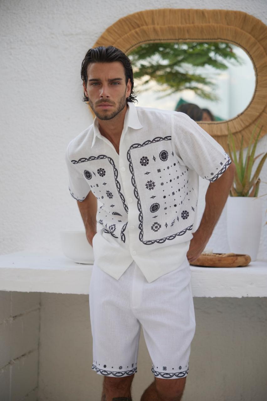 A man wearing a white shirt with a bandana pattern and white shorts standing in a room with a light-colored wall and a mirror.