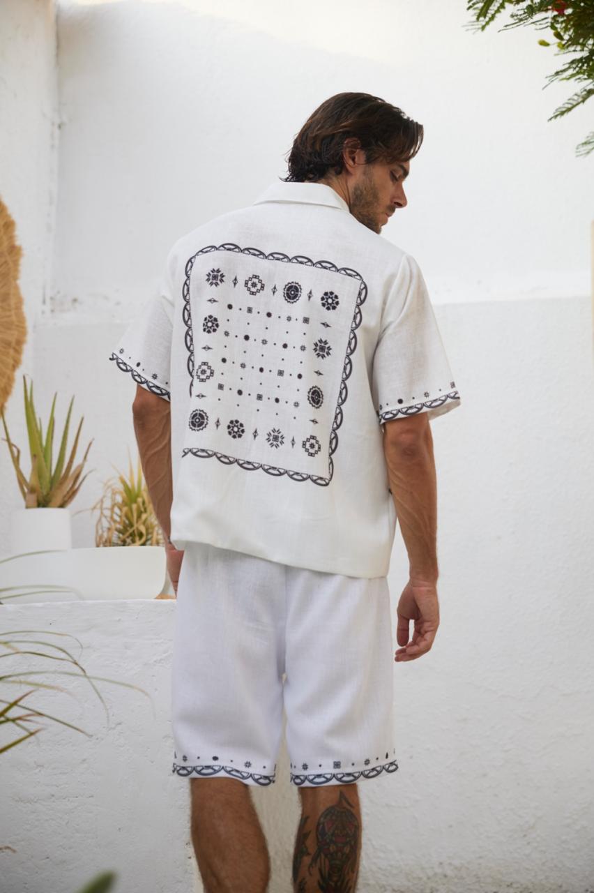 A man wearing a white shirt with a bandana pattern on the back and white shorts, standing against a white wall with plants in the background.