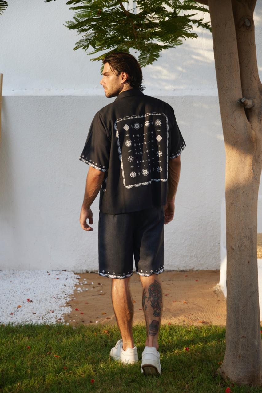 This image captures a stylish man from behind, wearing a unique, old-fashioned black short-sleeve shirt and matching shorts. The outfit features a distinctive floral pattern on the back, adding a vintage touch to his retro-inspired look.