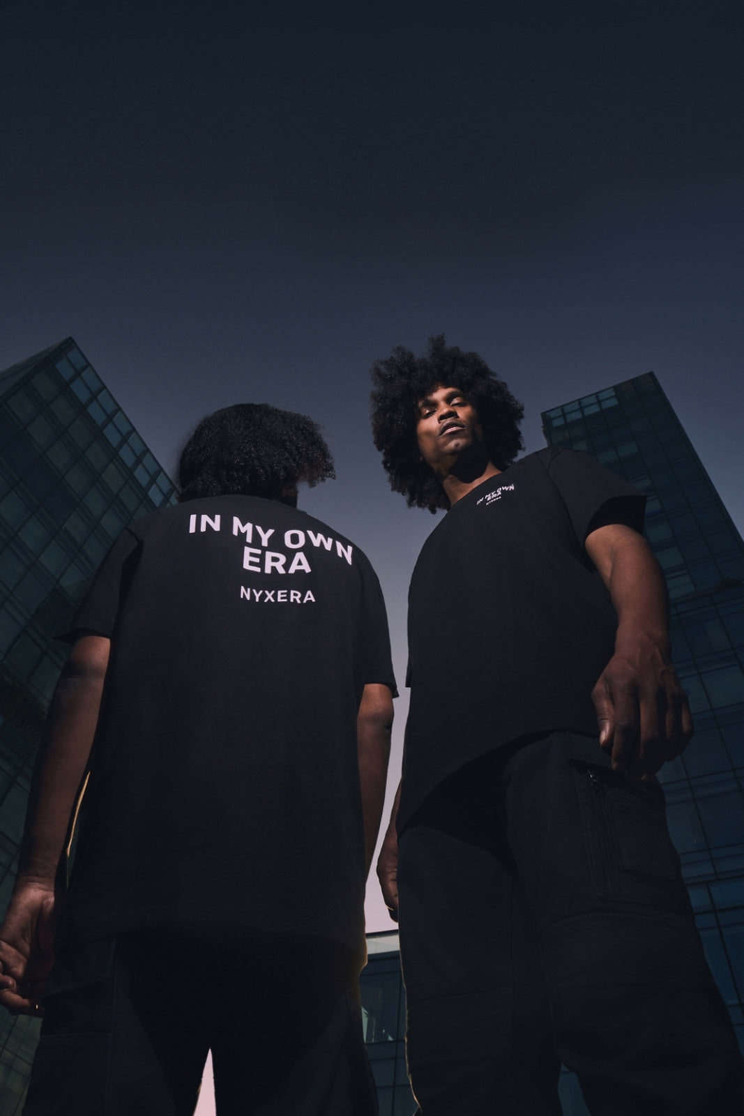 Two people wearing black t-shirts with text against a dark cityscape background