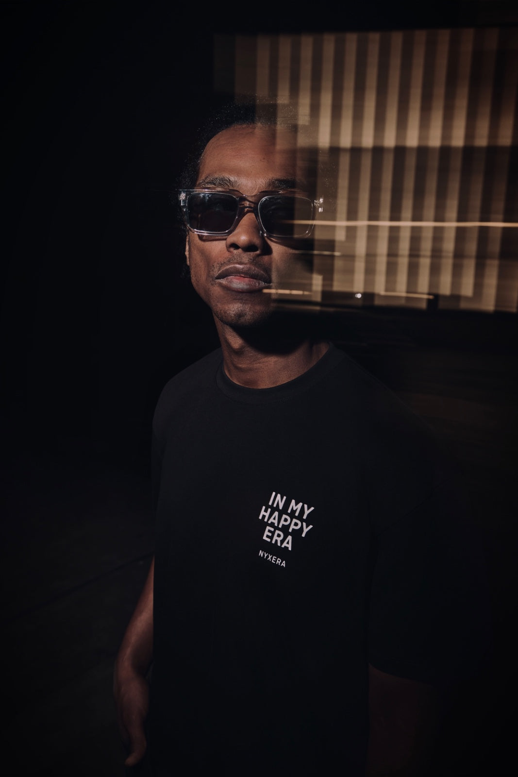 Person wearing sunglasses and a black t-shirt with text, standing against a dark background.