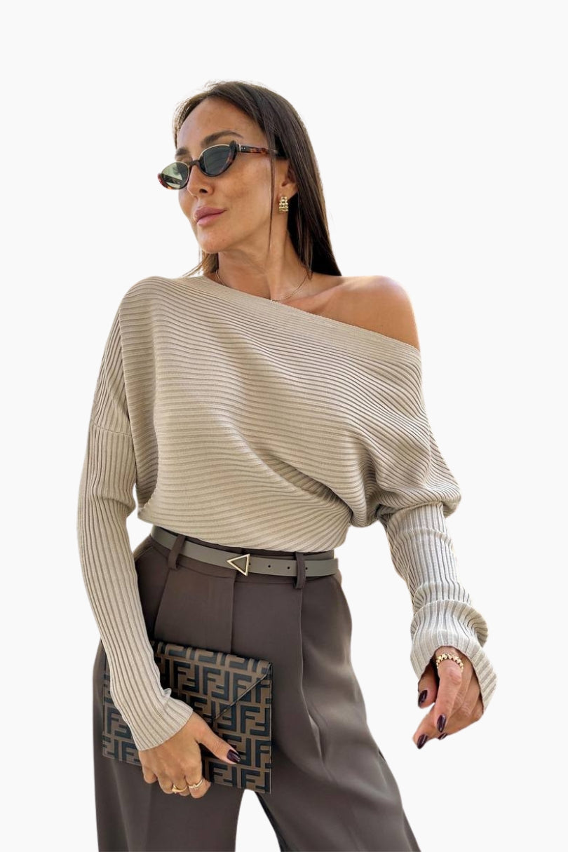 Woman wearing a beige off-shoulder sweater and gray pants, holding a patterned clutch.