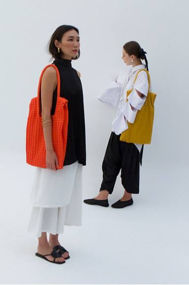 Two women standing side by side, each holding a large tote bag, one orange and one yellow, on a white background.