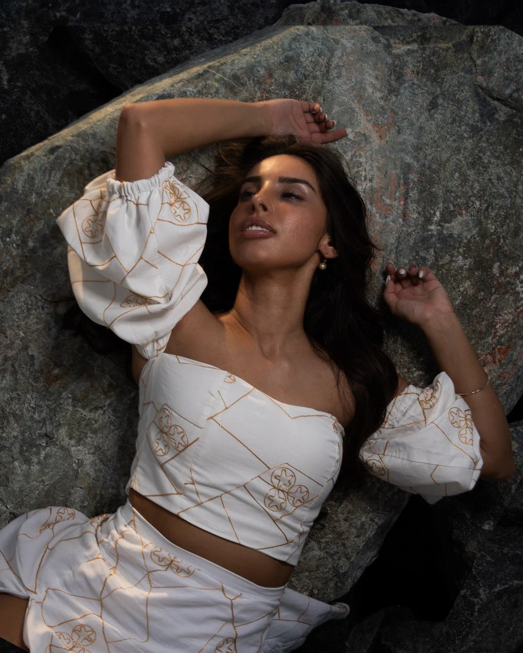 Woman in a white outfit with gold patterns lying on rocks. set is a white and gold patterned outfit. white cotton set, golden embroidery and voluminous sleeves. slit on the skirt.