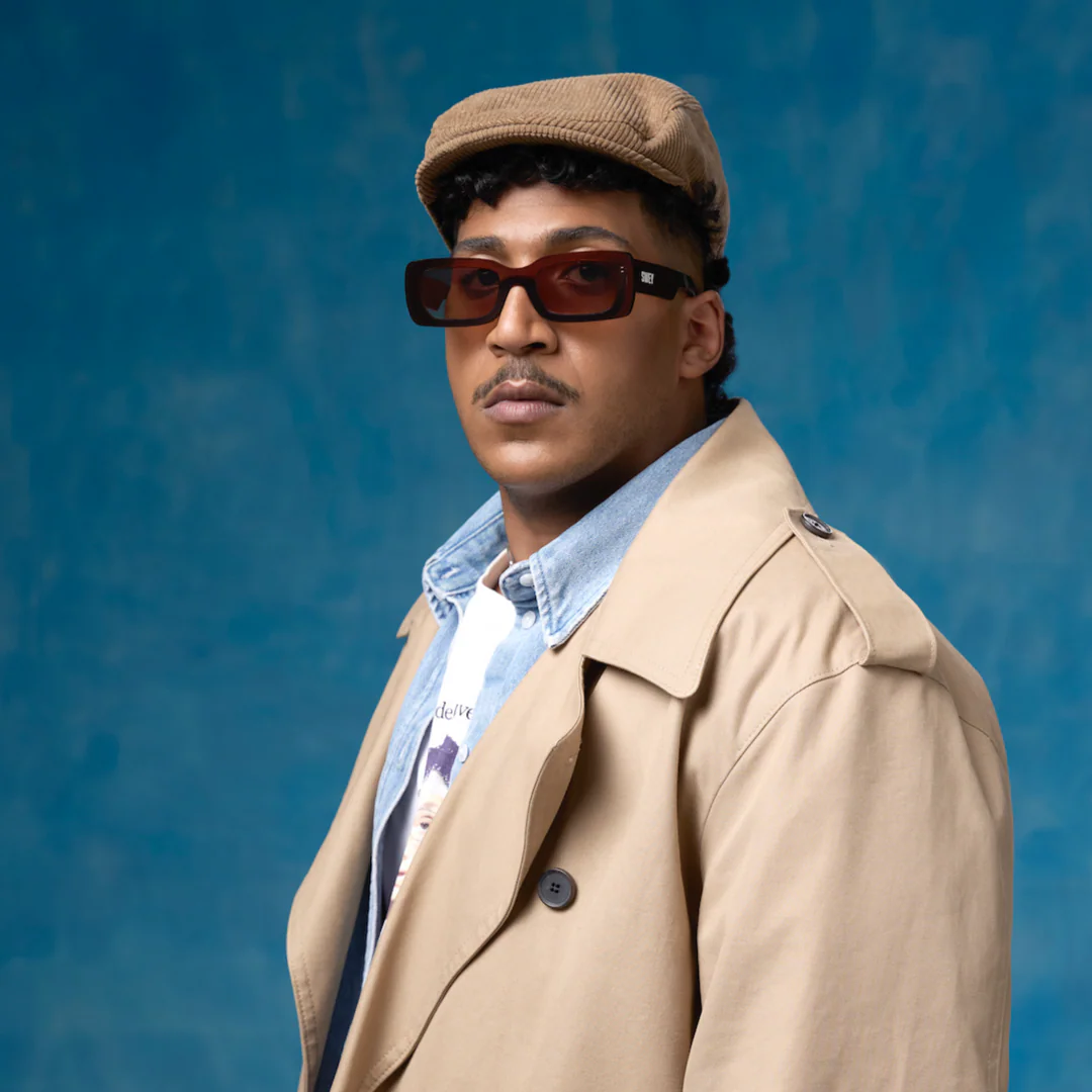 Man wearing a beige trench coat, cap, and sunglasses against a blue background