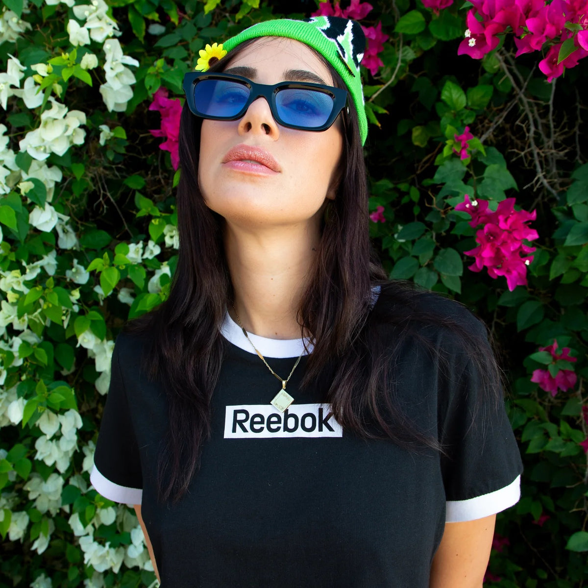 Person wearing a Reebok t-shirt with sunglasses and a green hat against a floral background