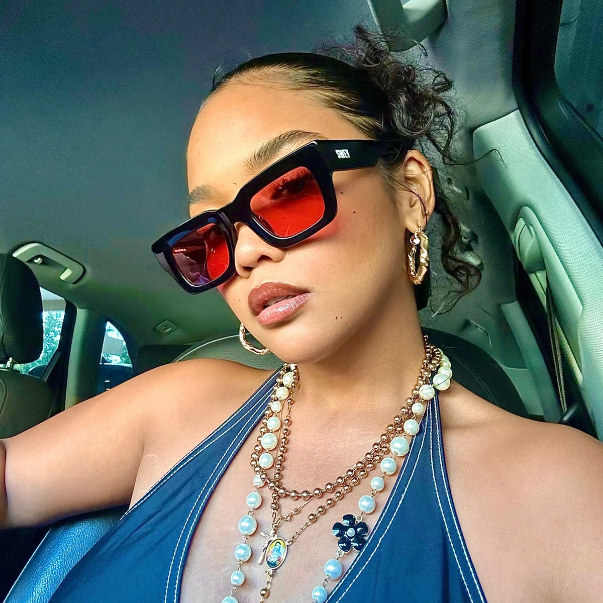 Woman wearing a black sunglass, pink lenses and a blue top inside a car