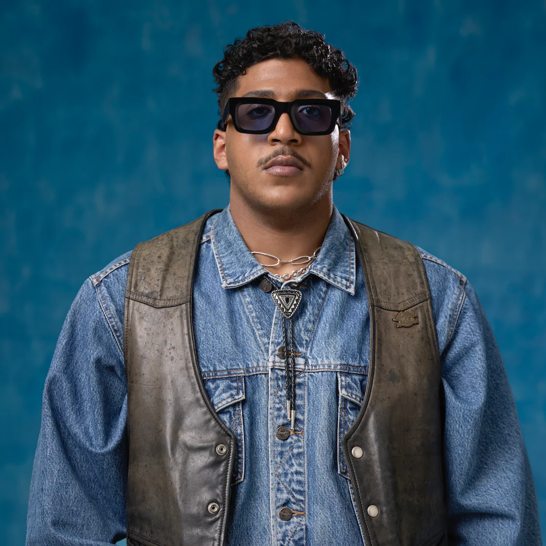 Man wearing sunglasses and a denim jacket with a leather vest against a blue background