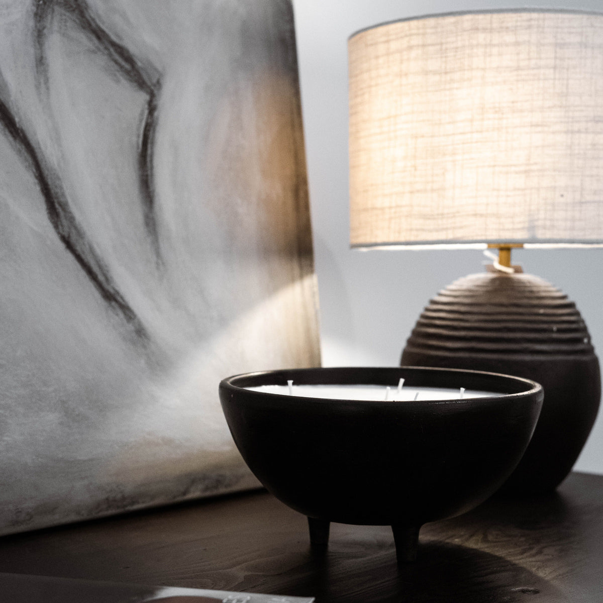 Candle in a black bowl on a wooden surface with a lamp and abstract painting in the background.