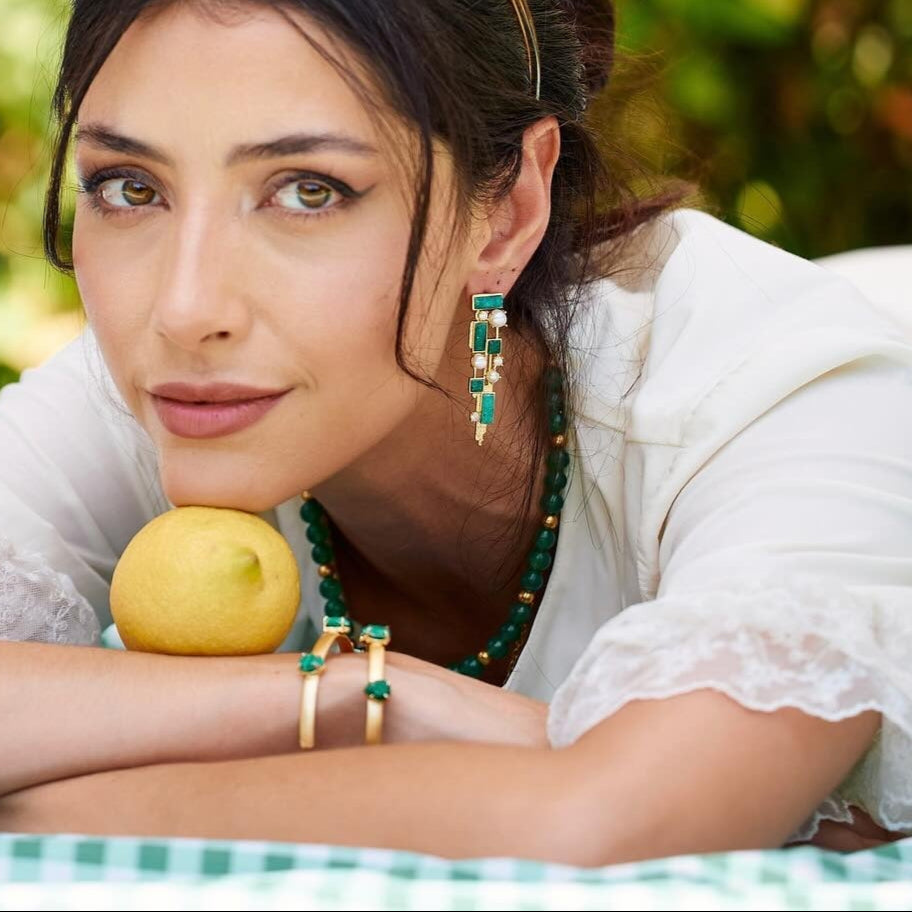 Woman with green earrings and necklace holding a lemon, wearing a white blouse.
