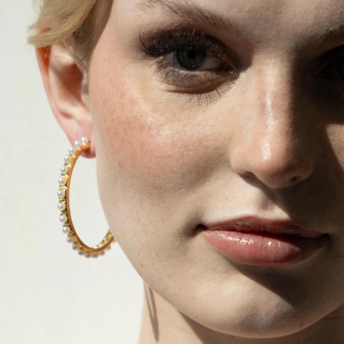 Close-up of a woman wearing gold hoop earrings and a necklace against a neutral background