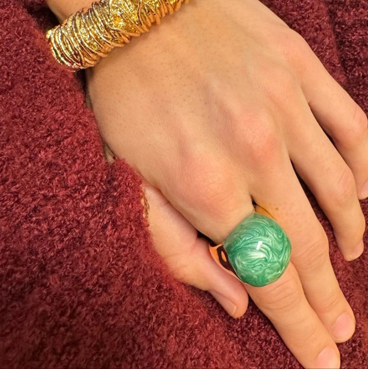 Hand wearing a gold bracelet and green ring on a red textured fabric background